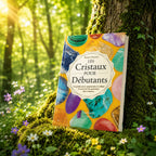 Livre Les Cristaux pour Débutants sur mousse sacrée au pied d'un arbre, entouré de fleurs violettes sous des rayons de soleil magiques.