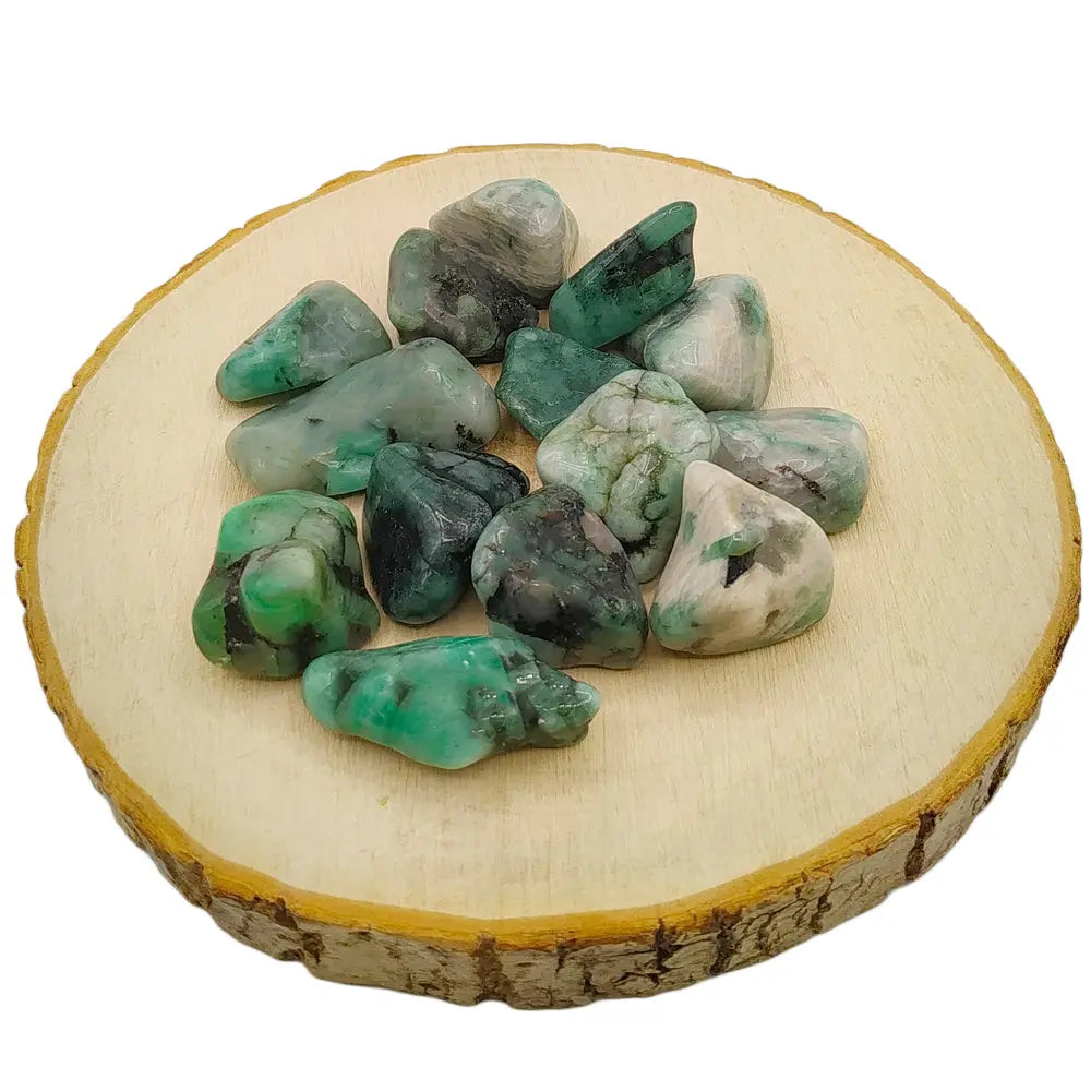 Un groupe de pierres d'émeraude roulées mêlant des nuances de vert vif à des inclusions naturelles de roche grise et noire, disposé sur une rondelle de bois clair.