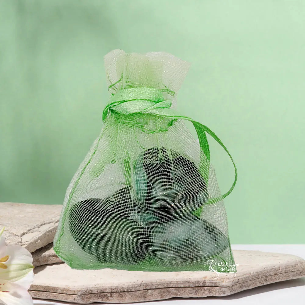 Un petit sac en organza vert contenant des pierres d'émeraude, posé sur une pierre plate naturelle devant un arrière-plan vert clair.