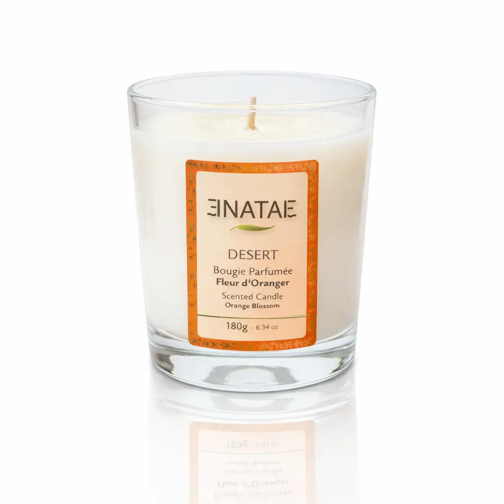 Une bougie parfumée naturelle « Hawaïki » (Monoï - Coco) de la marque Enatae, présentée dans un verre transparent simple, sur un fond blanc avec un reflet à sa base.