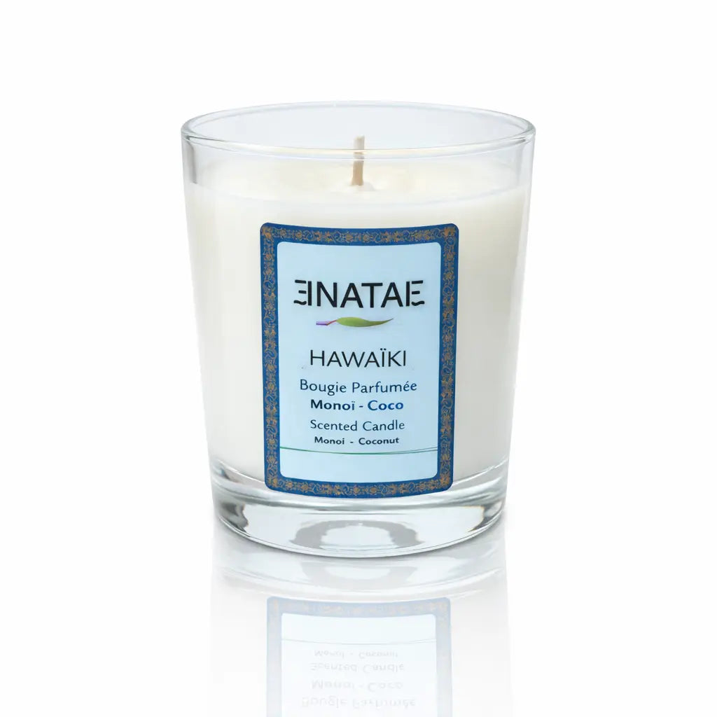 Une bougie parfumée naturelle « Hawaïki » (Monoï - Coco) de la marque Enatae, présentée dans un verre transparent simple, sur un fond blanc avec un reflet à sa base.