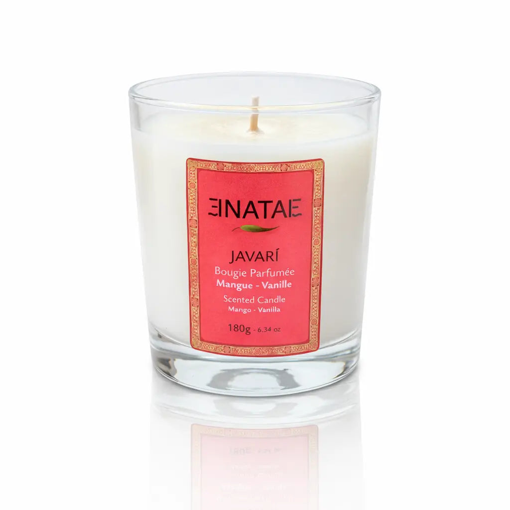 Une bougie parfumée naturelle « Javari » (Mangue - Vanille) de la marque Enatae, présentée dans un verre transparent simple, sur un fond blanc avec un reflet à sa base.