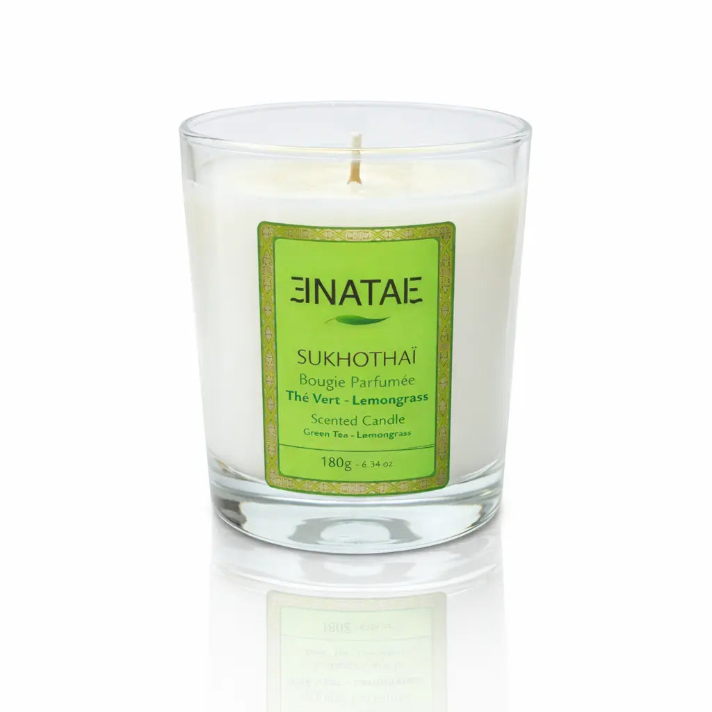 Une bougie parfumée naturelle « Sukhothaï » (Thé Vert - Lemongrass) de la marque Enatae, présentée dans un verre transparent simple, sur un fond blanc avec un reflet à sa base.