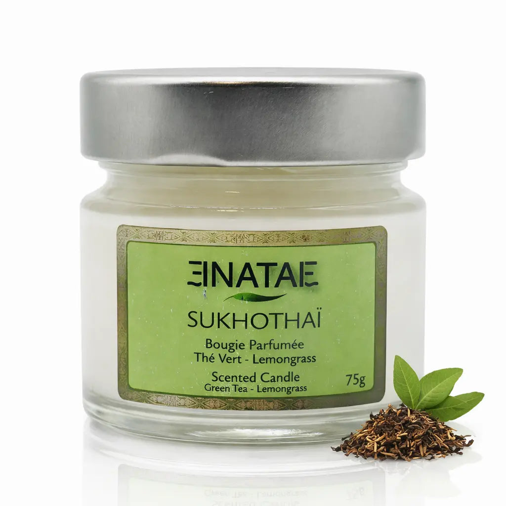 Une bougie parfumée naturelle « Sukhothaï » (Thé Vert - Lemongrass) de la marque Enatae, présentée dans un petit pot en verre avec couvercle métallique, accompagnée d'un petit tas de thé et de feuilles sur un fond blanc.