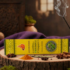 Encens Ayurvedic Patchouli en étui rectangulaire jaune vif posé sur une tranche de bois, entouré d'épices, de bâtons de cannelle et d'un bâtonnet d'encens fumant.