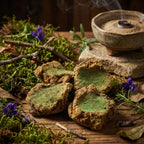 Morceaux d’encens verts et bruns disposés sur table en bois, avec porte-encens en céramique fumant, mousse, feuilles et fleurs violettes.