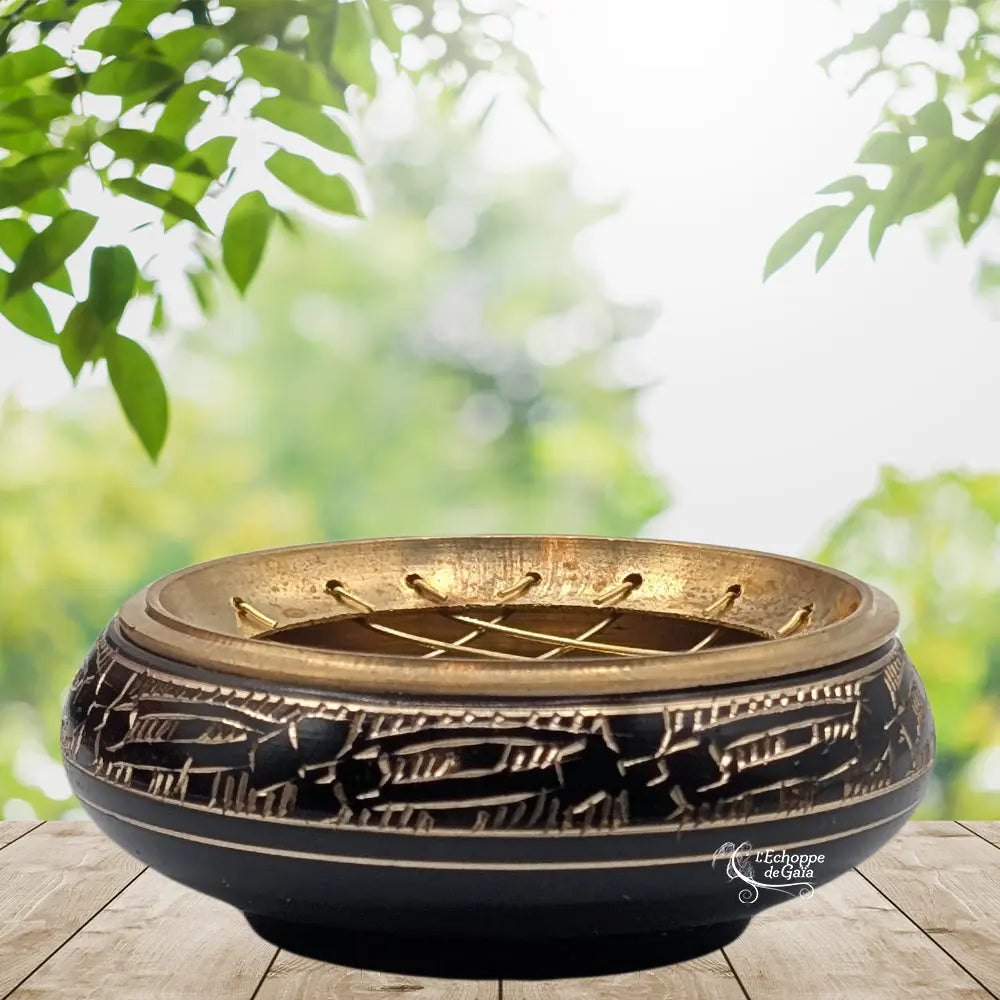 Brûle-encens bas en métal noir et doré, motifs géométriques gravés sur le pourtour, grille croisée, présenté sur planche en bois, décor végétal.