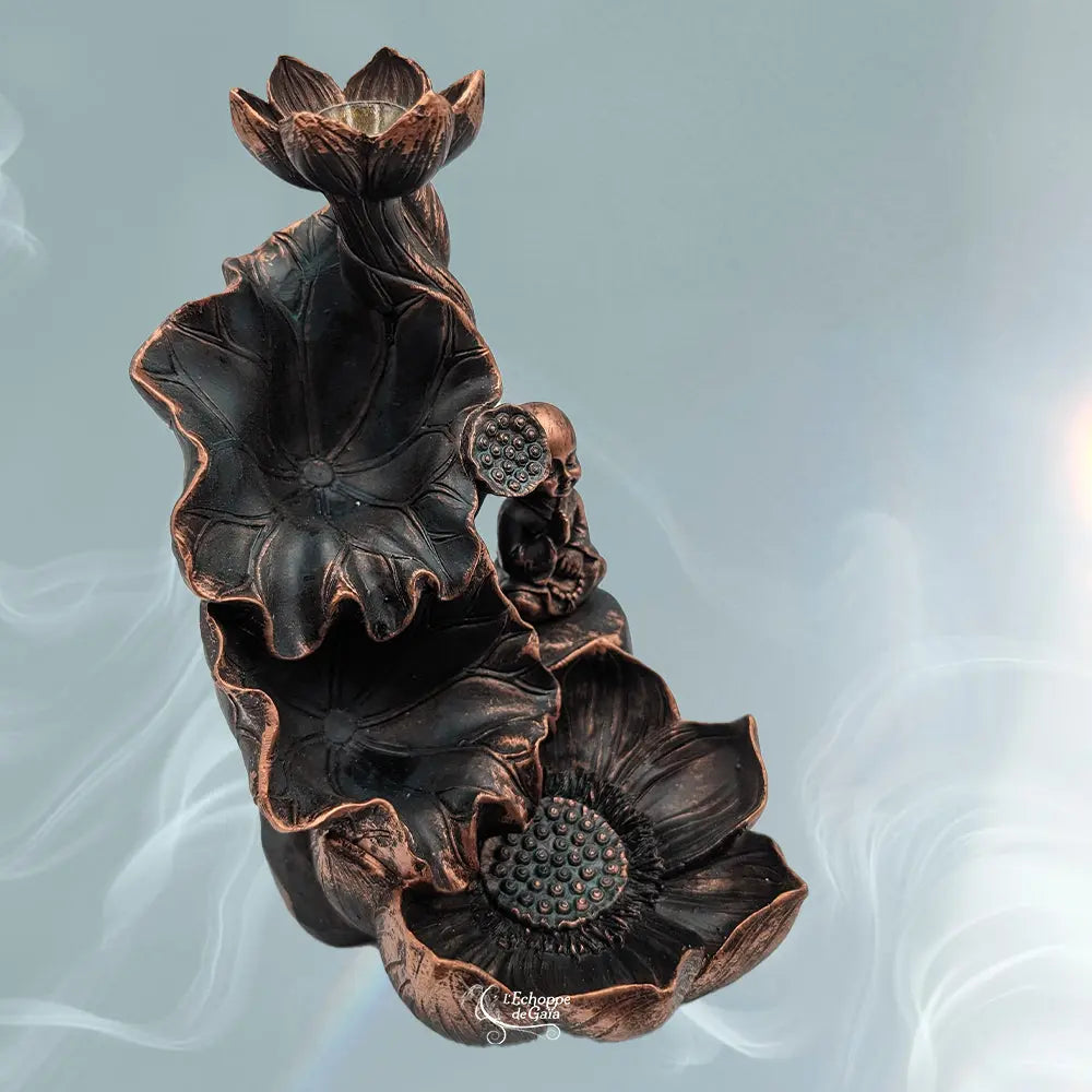 Même fontaine à encens backflow, vue plus “de dessus” : trois niveaux (fleur/lotus en haut, grande feuille en cuvette au milieu, grande fleur/coupelle en bas), avec le petit bouddha assis à droite. Finition noir-bronze/cuivrée, fond bleu-gris avec volutes de fumée.