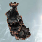 Même fontaine à encens backflow, vue plus “de dessus” : trois niveaux (fleur/lotus en haut, grande feuille en cuvette au milieu, grande fleur/coupelle en bas), avec le petit bouddha assis à droite. Finition noir-bronze/cuivrée, fond bleu-gris avec volutes de fumée.