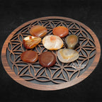 Une collection de galets de cornaline polis, aux motifs et nuances variés (orangé, rouge, translucide), disposés sur une fleur de vie en bois sculpté.