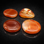 Quatre galets de cornaline polis aux teintes rouge orangé intense, présentant des surfaces lisses et brillantes avec des bandes de couleur naturelles, disposés sur un support noir sombre.
