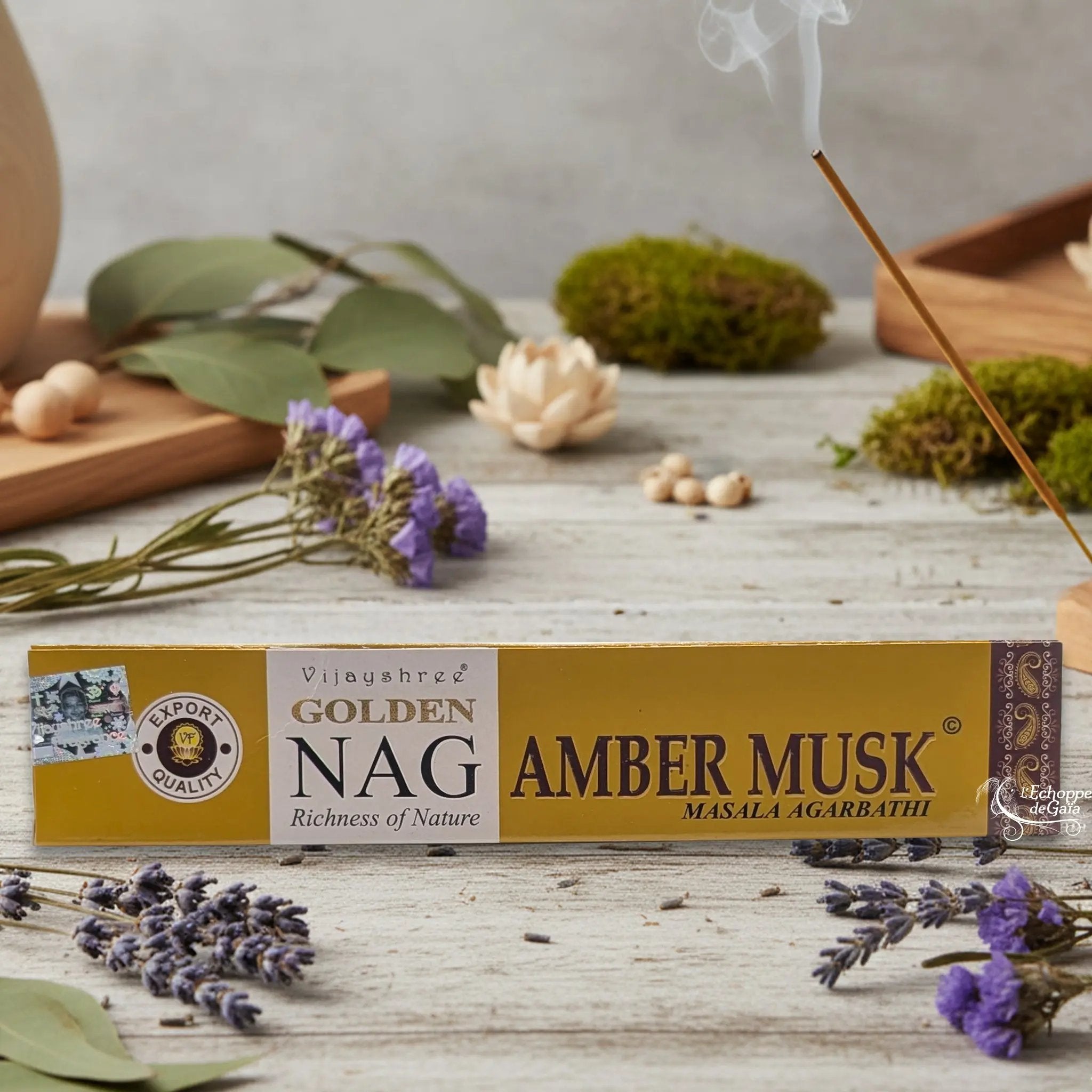 Boîte d'encens Vijayshree Golden Nag "Amber Musk" Masala Agarbathi, posée sur une table avec de la lavande et un bâton d'encens fumant.