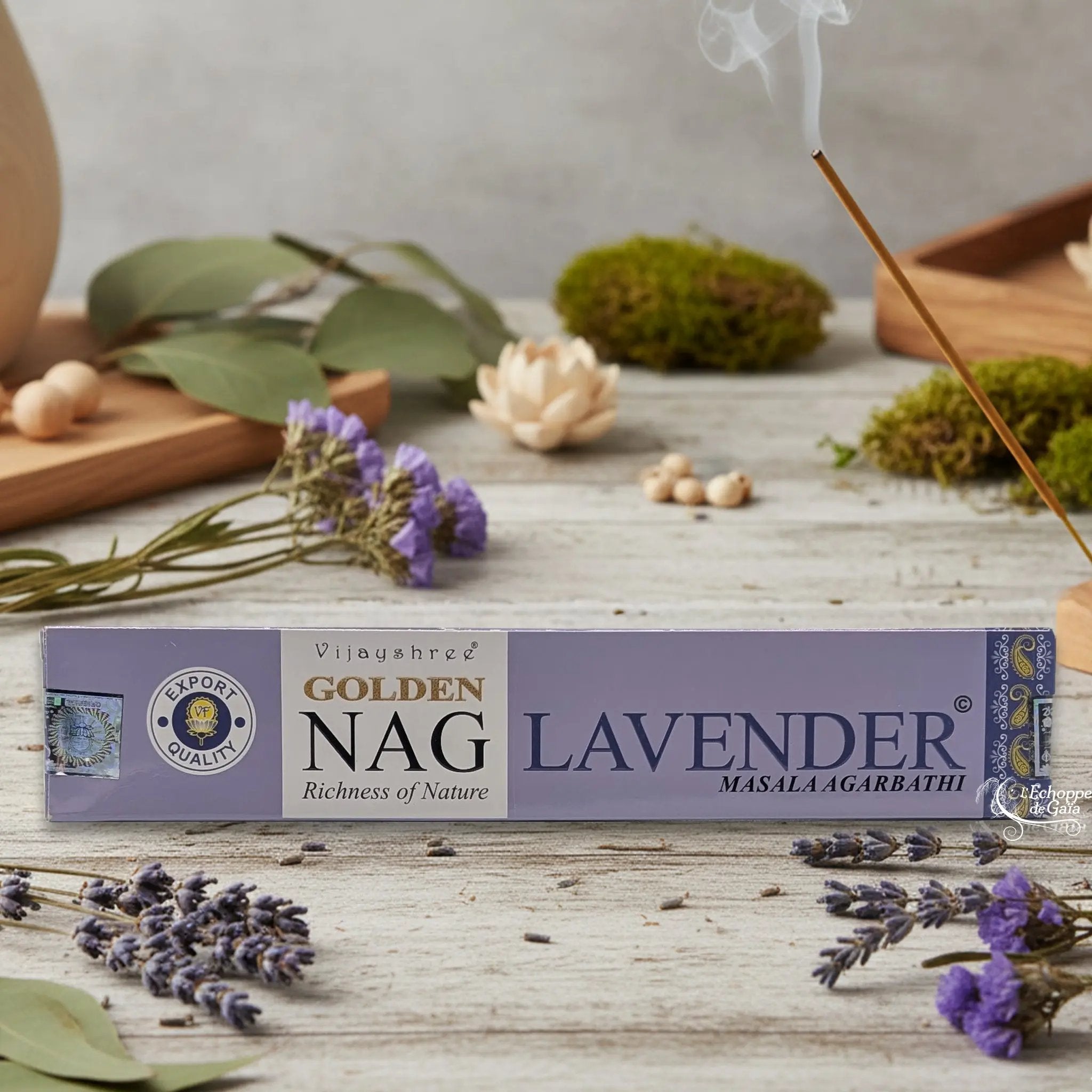 Boîte d'encens Vijayshree Golden Nag "Lavender" Masala Agarbathi, posée sur une table avec de la lavande et un bâton d'encens fumant.