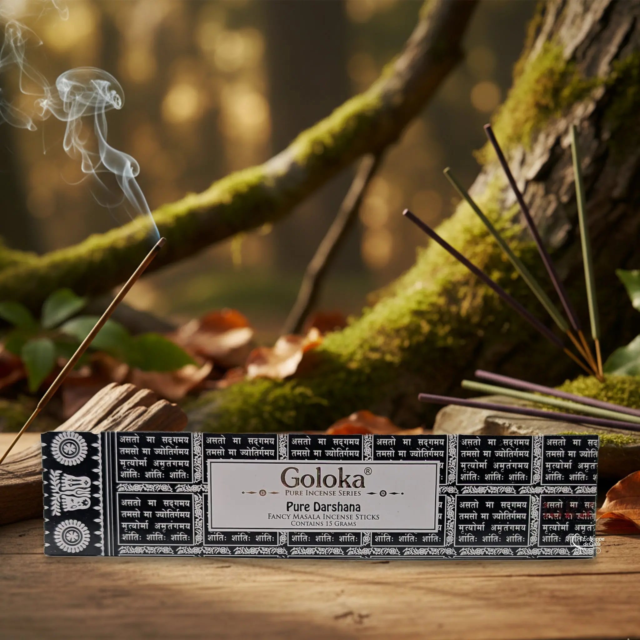 Boîte d’encens Goloka « Pure Darshana » posée sur une table en bois, avec un bâtonnet allumé dégageant de la fumée, sur fond de forêt moussue.