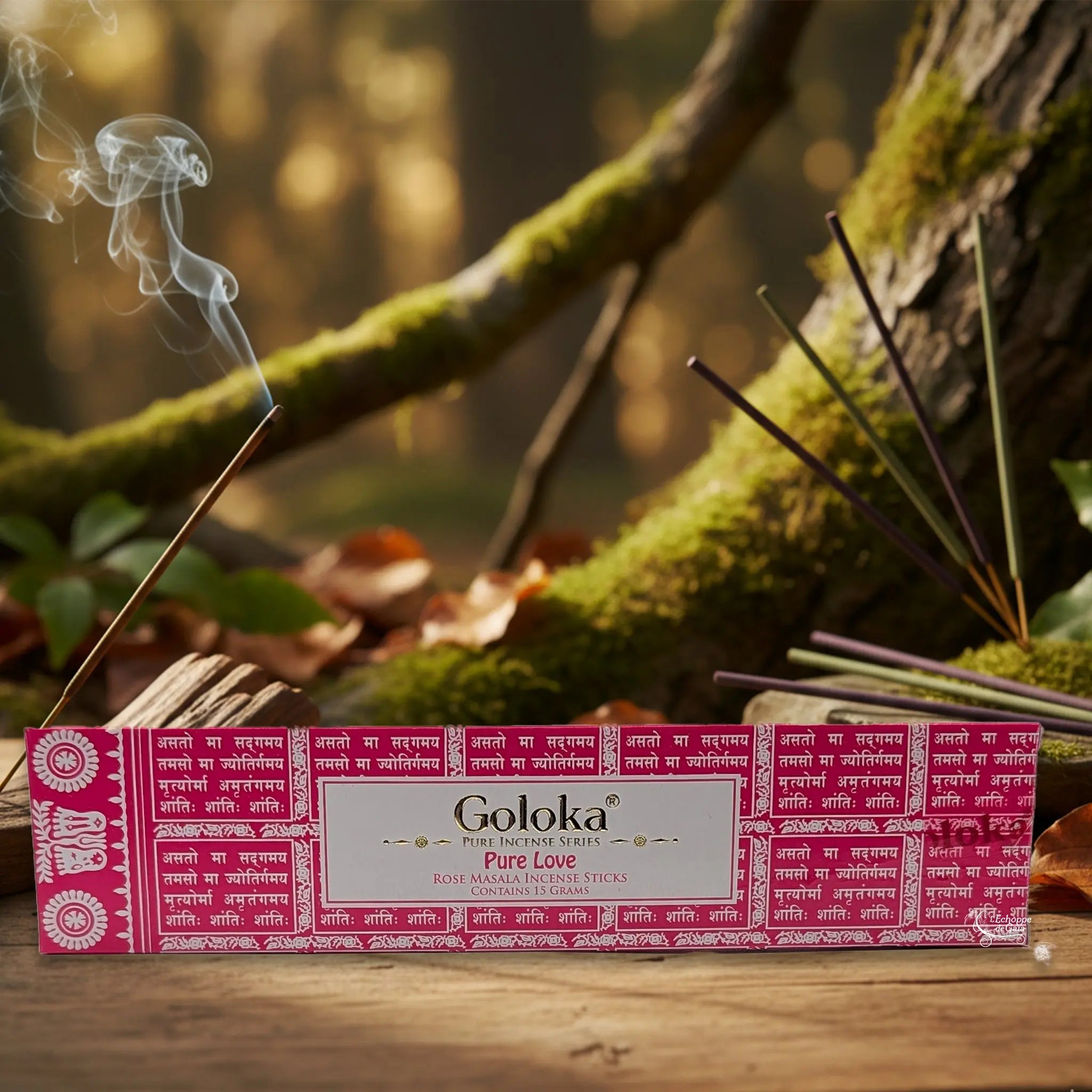 Boîte d’encens Goloka « Pure Love » (rose) posée sur une table en bois, avec un bâtonnet allumé dégageant de la fumée, sur fond de forêt moussue.