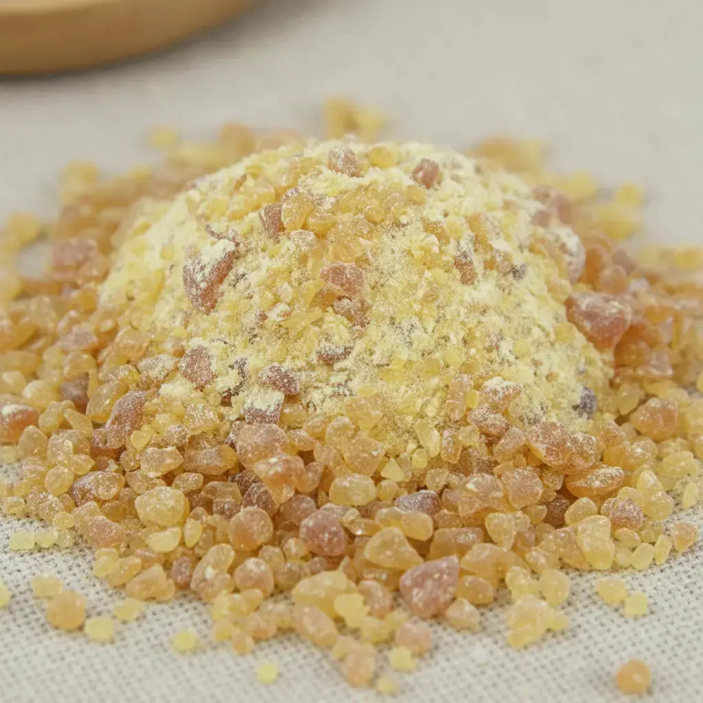 Gros plan sur un monticule de grains de résine d'Élémi, montrant de l'ambre clair mélangé à de la poudre de résine jaune pâle, sur un fond en tissu clair.