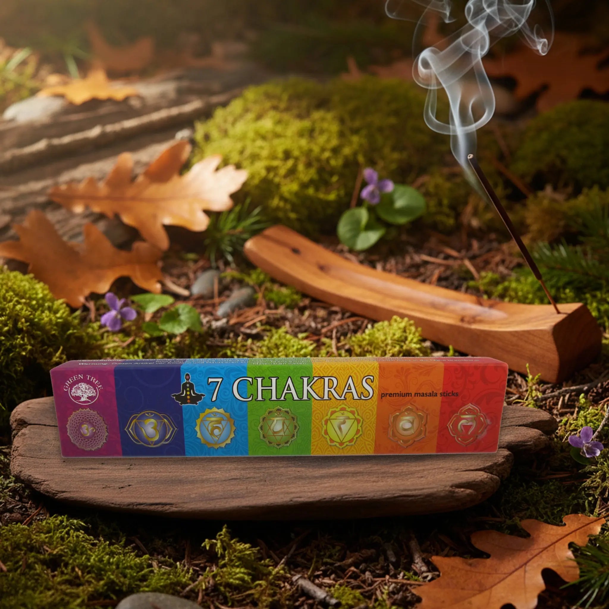 Boîte d’encens Green Tree “7 Chakras” posée sur un support en bois, avec bâton d’encens fumant en arrière-plan sur sol forestier.