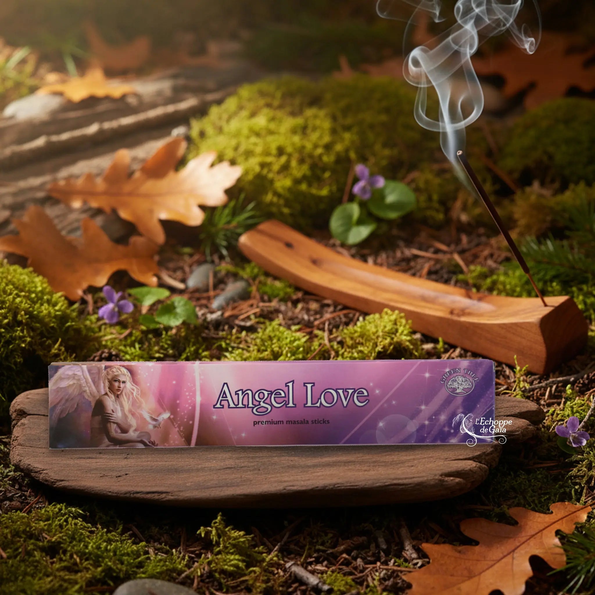 Boîte d’encens Green Tree “Angel Love” en ton rose/violet, posée sur une planche de bois au milieu de la mousse et des feuilles d’automne, avec un porte-encens en bois fumant à l’arrière-plan.