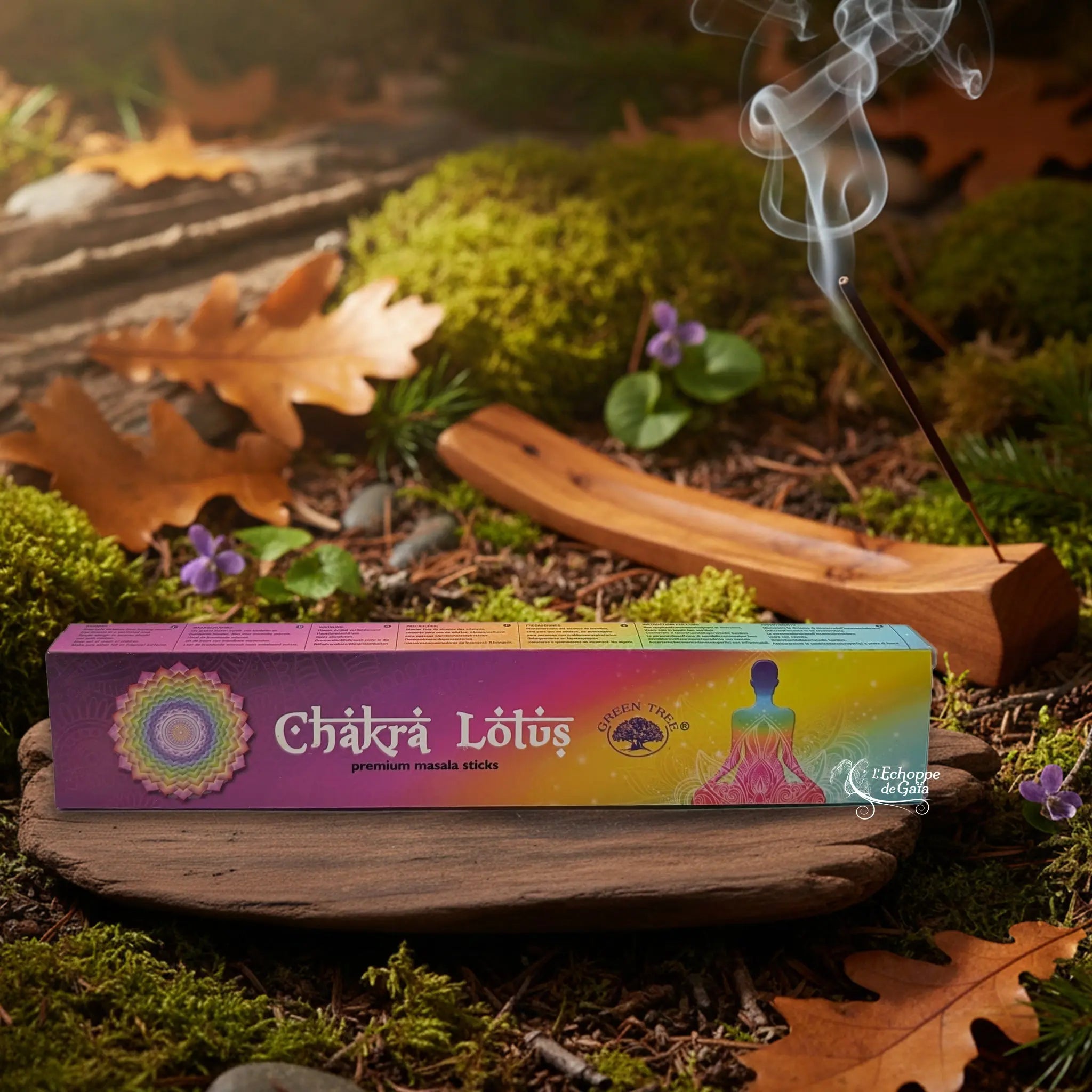 Boîte d’encens Green Tree “Chakra Lotus” aux couleurs arc-en-ciel (violet, rose, jaune, vert, bleu), avec un mandala à gauche et la silhouette d’une personne en méditation sur un lotus à droite, posée sur une planche de bois au milieu de mousse et de feuilles, un bâton d’encens fumant dans un porte-encens en bois à l’arrière-plan.