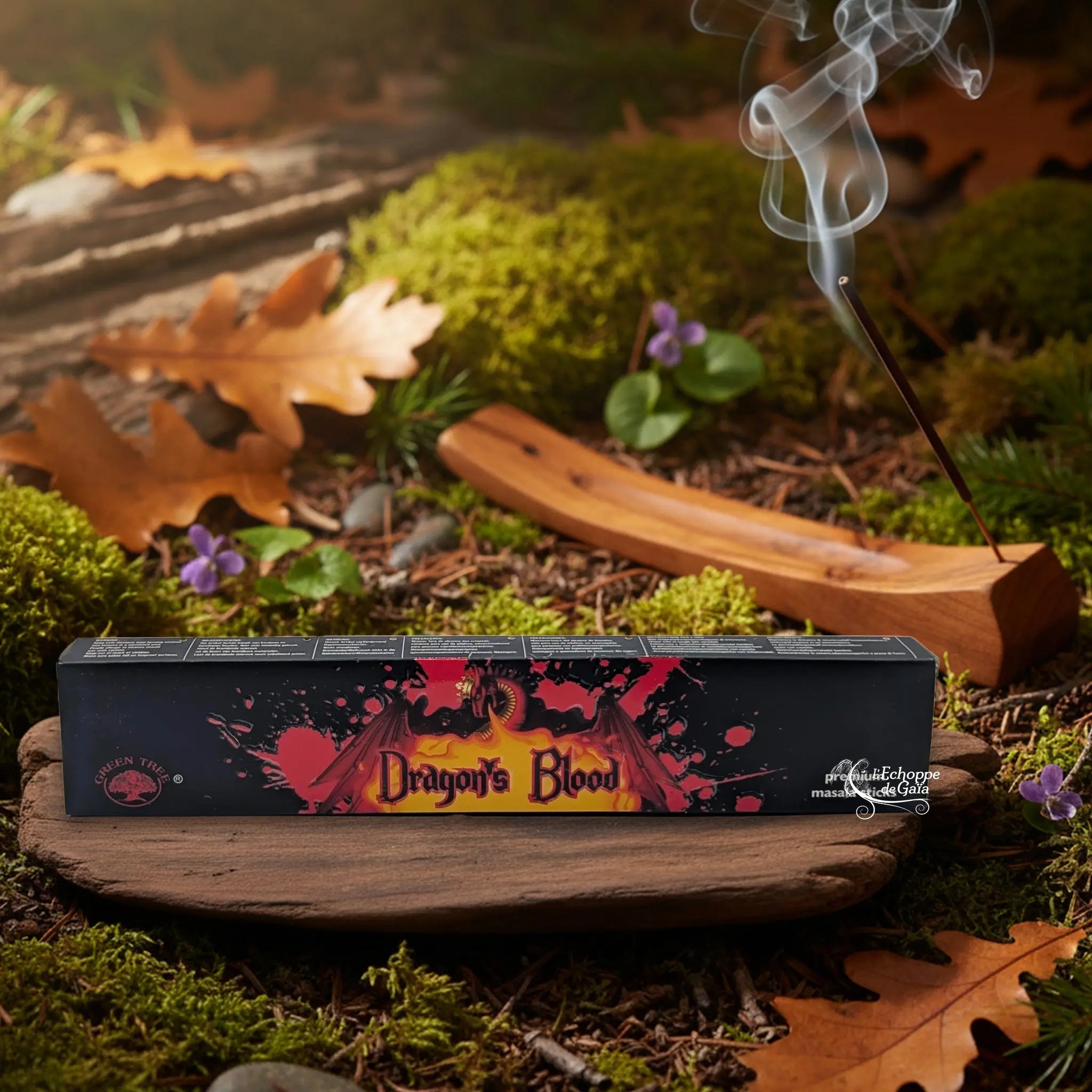 Boîte d’encens Green Tree “Dragon’s Blood” posée sur un support en bois, avec bâton d’encens fumant en arrière-plan sur sol forestier.
