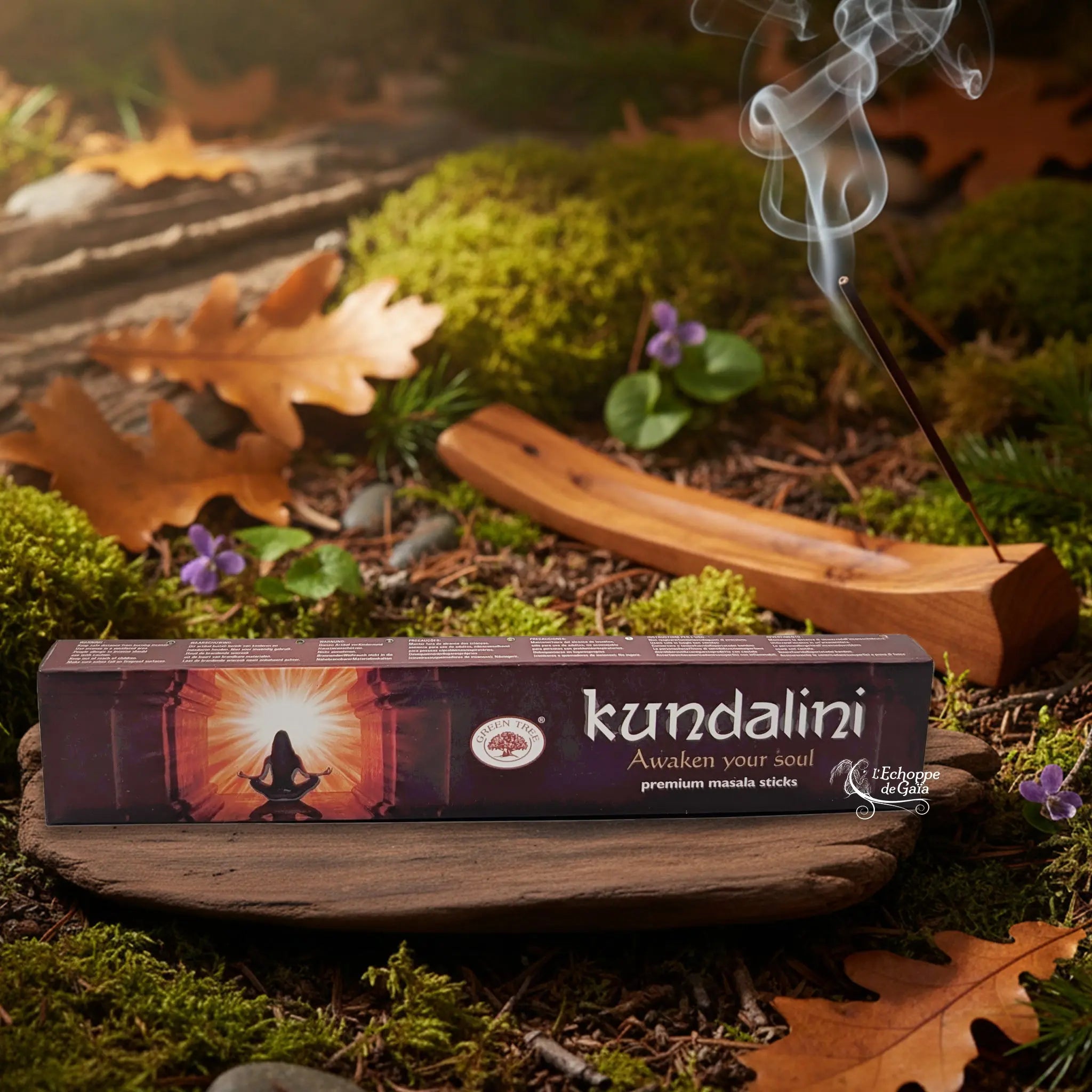 Boîte d’encens Green Tree “Kundalini” posée sur un support en bois, avec bâton d’encens fumant en arrière-plan sur sol forestier.
