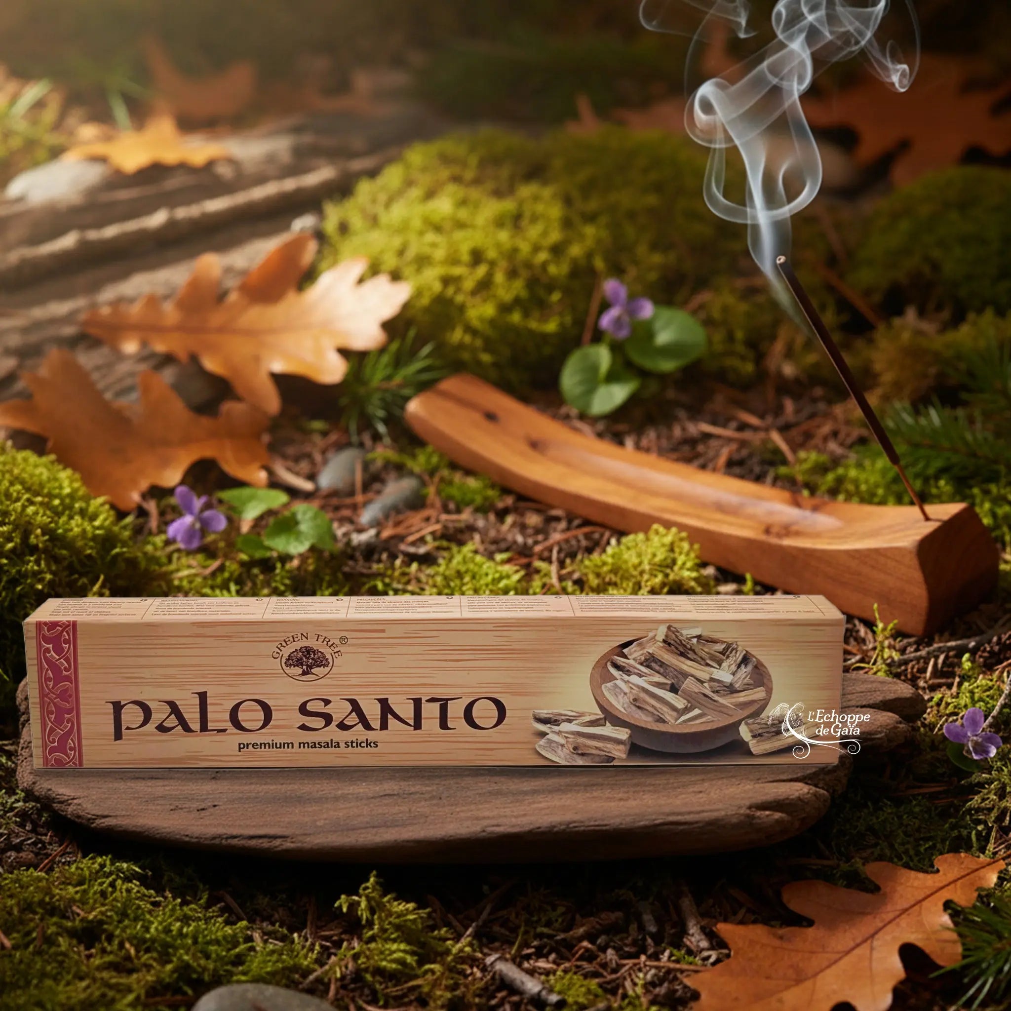 Boîte d’encens Palo Santo Green Tree posée sur un support en bois, avec bâton d’encens fumant en arrière-plan sur un sol forestier.
