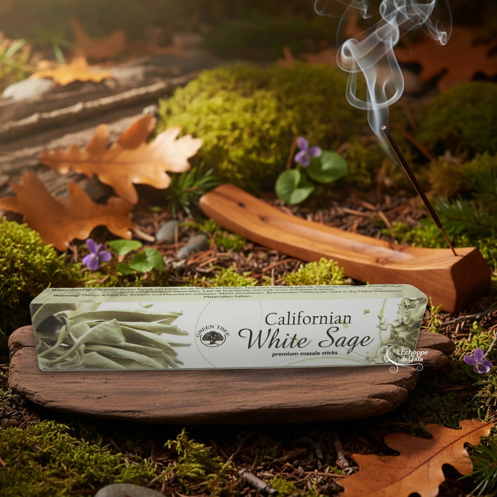Boîte d’encens Green Tree “Californian White Sage” posée sur un support en bois, avec bâton d’encens fumant en arrière-plan sur sol forestier.
