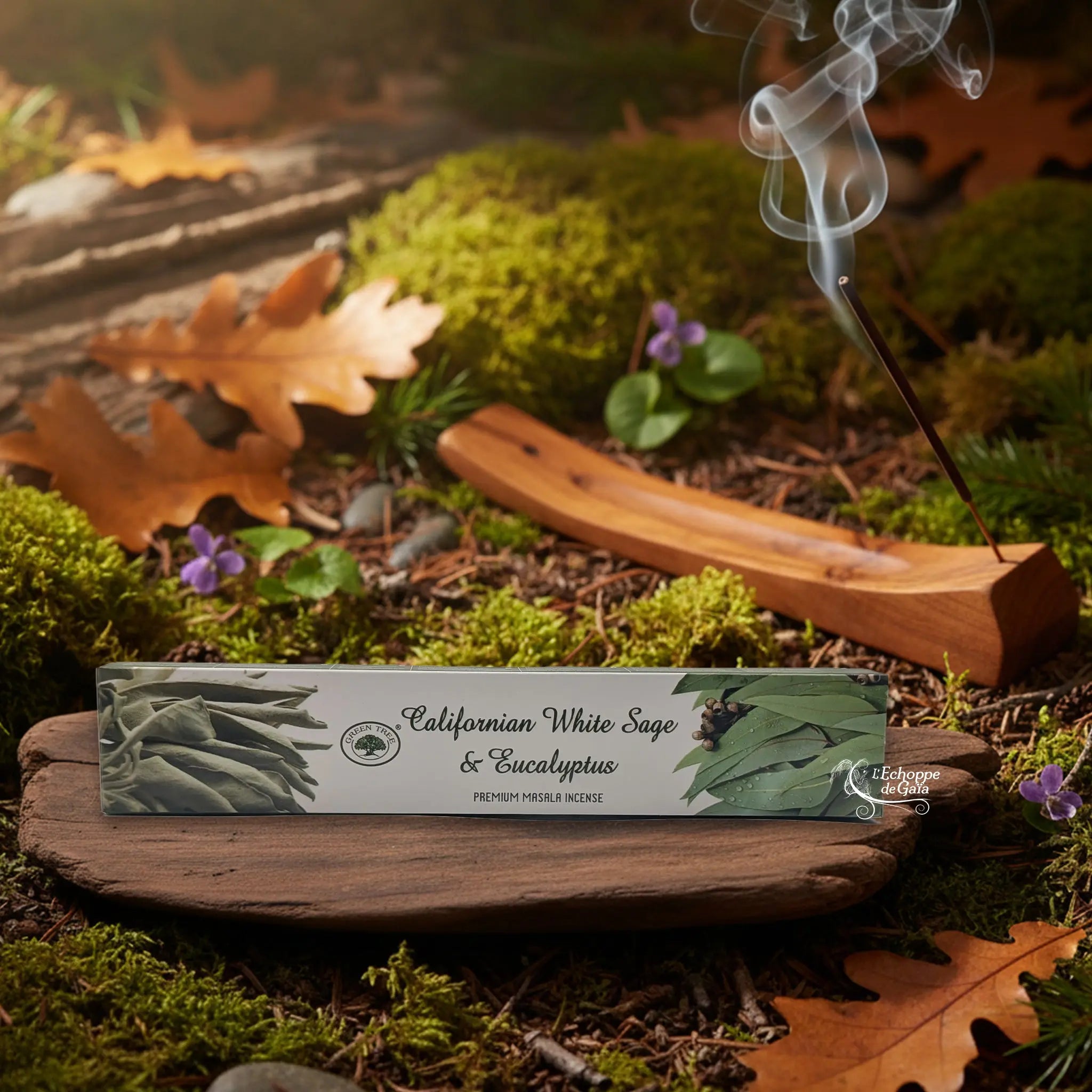 Boîte d’encens Green Tree « Californian White Sage & Eucalyptus » posée sur une tranche de bois, avec un bâtonnet allumé dans un porte-encens en bois et de la fumée, décor nature.