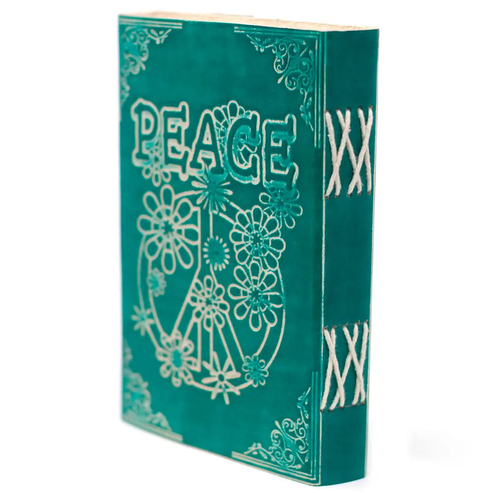 La couverture d'un carnet grimoire en cuir turquoise présentant le mot "PEACE" et un symbole de paix fleuri embossés, avec une reliure artisanale à lacets blancs apparents sur le dos.