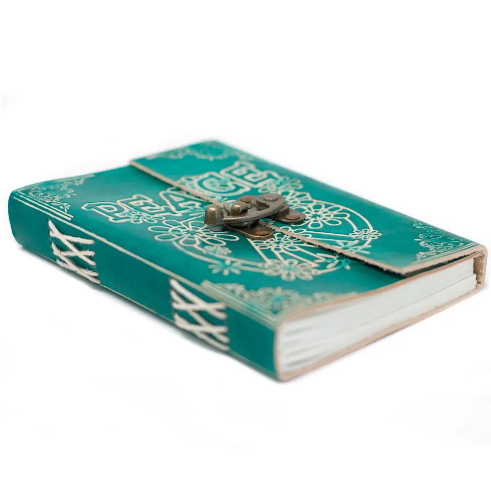 Un carnet grimoire en cuir turquoise embossé du mot "PEACE" et de motifs floraux, fermé par un loquet métallique en laiton et relié par des surpiqûres blanches apparentes sur fond blanc.