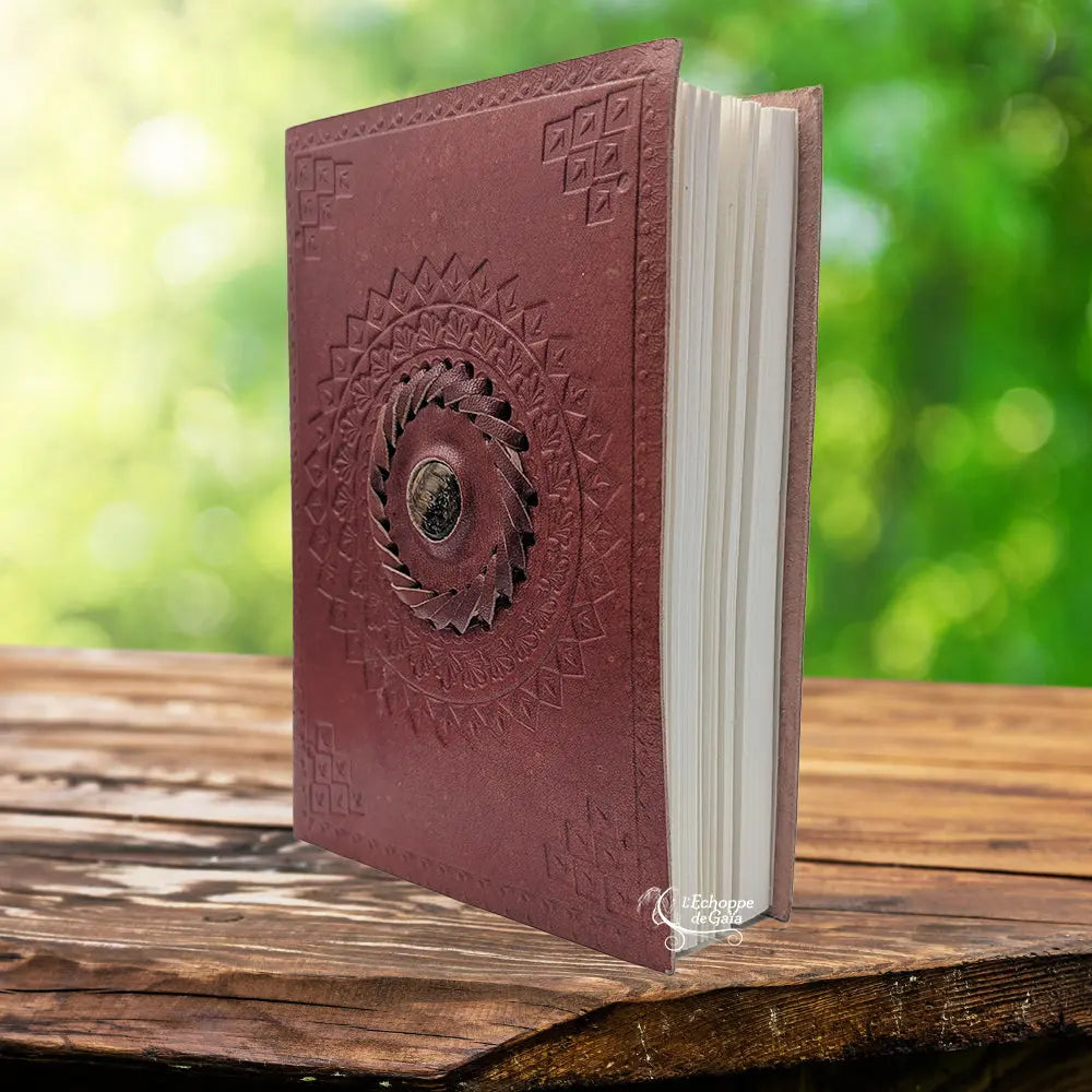 Un carnet grimoire en cuir marron clair entrouvert, orné d'une pierre d'œil de tigre centrale sertie dans une tresse de cuir et entourée de mandalas embossés, présenté sur un support en bois.