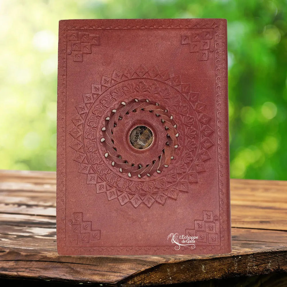 La couverture d'un carnet grimoire en cuir marron clair ornée d'une pierre d'œil de tigre centrale sertie dans une tresse de cuir, entourée de motifs mandalas et d'angles embossés, présentée sur un support en bois.