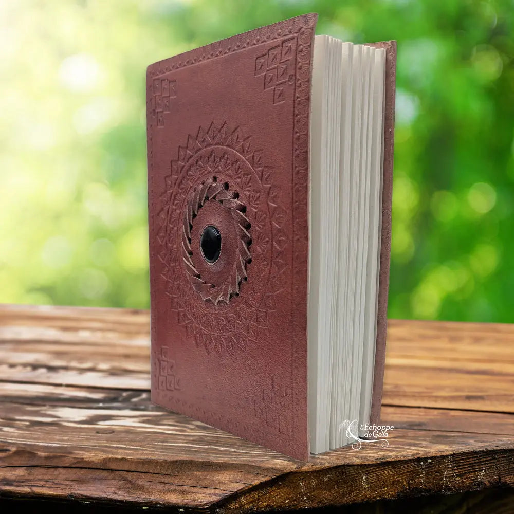 Un carnet grimoire en cuir marron clair entrouvert, orné d'une pierre d'onyx noire centrale sertie dans une tresse de cuir et entourée de mandalas embossés, présenté sur un support en bois.