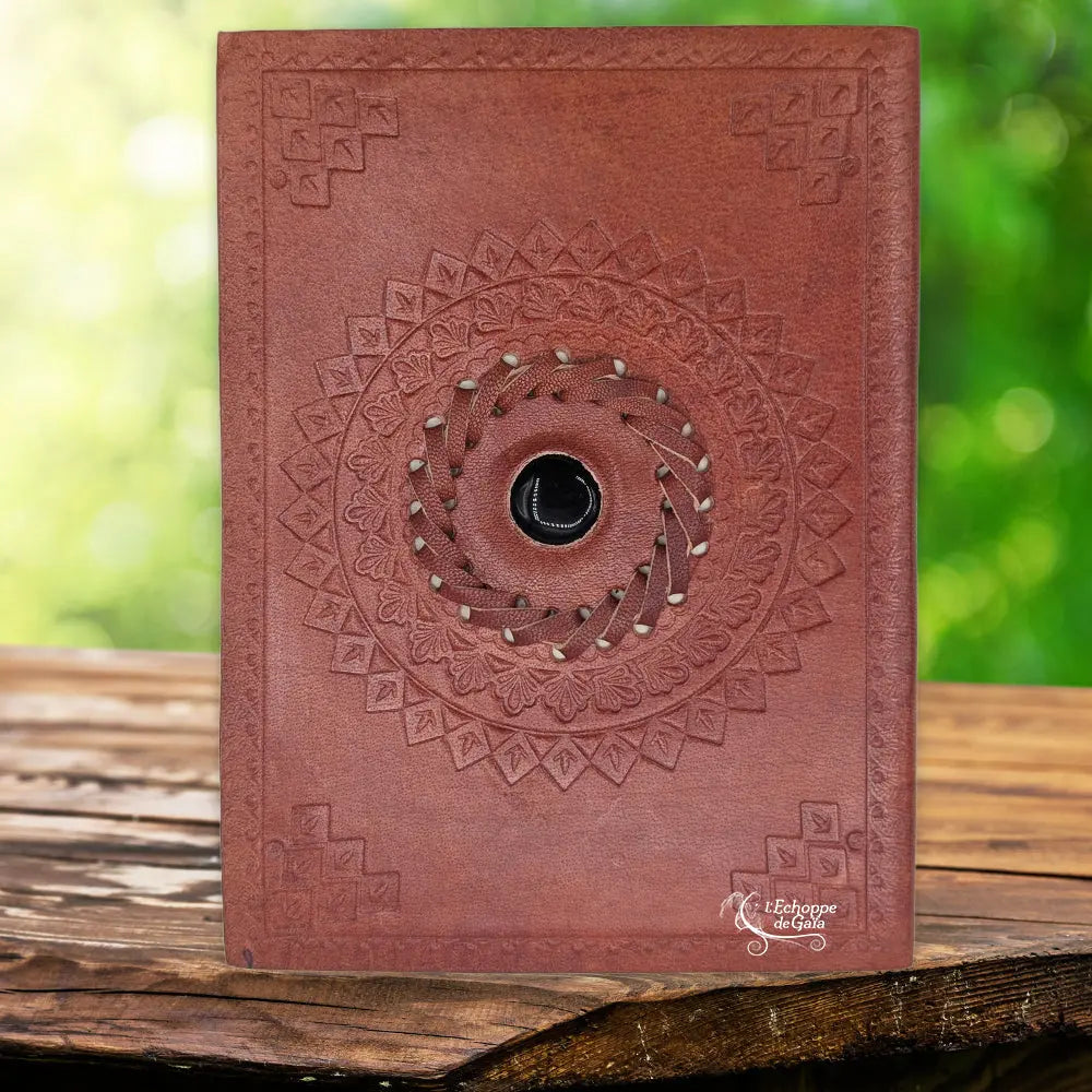 La couverture d'un carnet grimoire en cuir marron clair ornée d'une pierre d'onyx noire centrale sertie dans une tresse de cuir, entourée de motifs mandalas et d'angles embossés, présentée sur un support en bois.