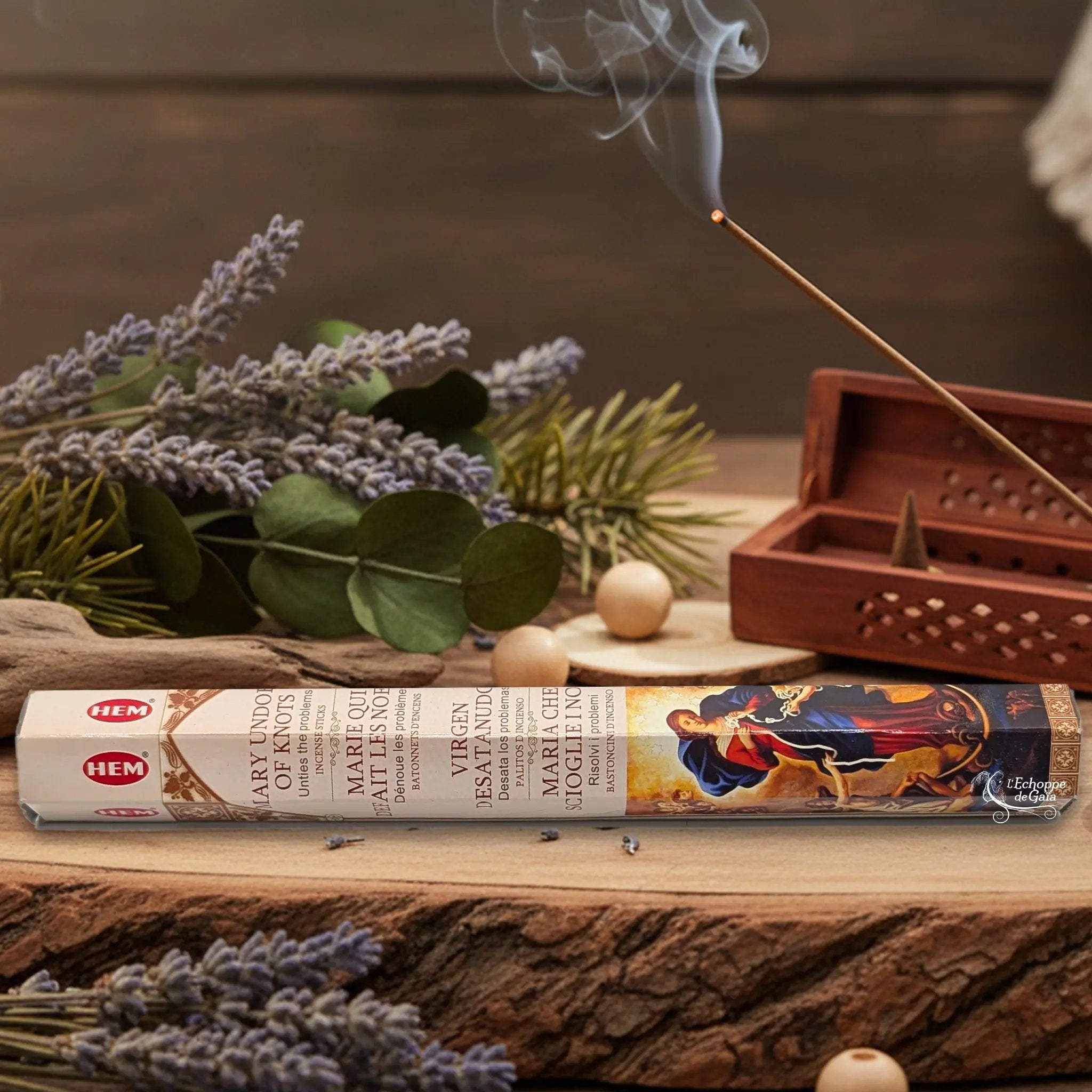 Boîte d'encens HEM "Marie qui défait les noeuds" posée sur du bois avec de la lavande et un bâton d'encens fumant.