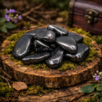 Pierres roulées d'Hématite aux reflets métalliques gris anthracite, luisantes sur une tranche de bois moussue, évoquant un trésor minéral protégé par la forêt.