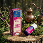 Flacon en verre bleu et son étui Herbes et Traditions avec une étiquette rose, posés sur une souche de bois entourée de mousse, de feuillages et d'un alambic en cuivre en arrière plan.