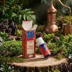 Flacon en verre bleu et son étui Herbes et Traditions - Huiles Essentielles Bio à Diffuser - Magie de Noël posés sur une souche de bois avec de la mousse, des pommes de pin et un alambic en cuivre en arrière-plan.