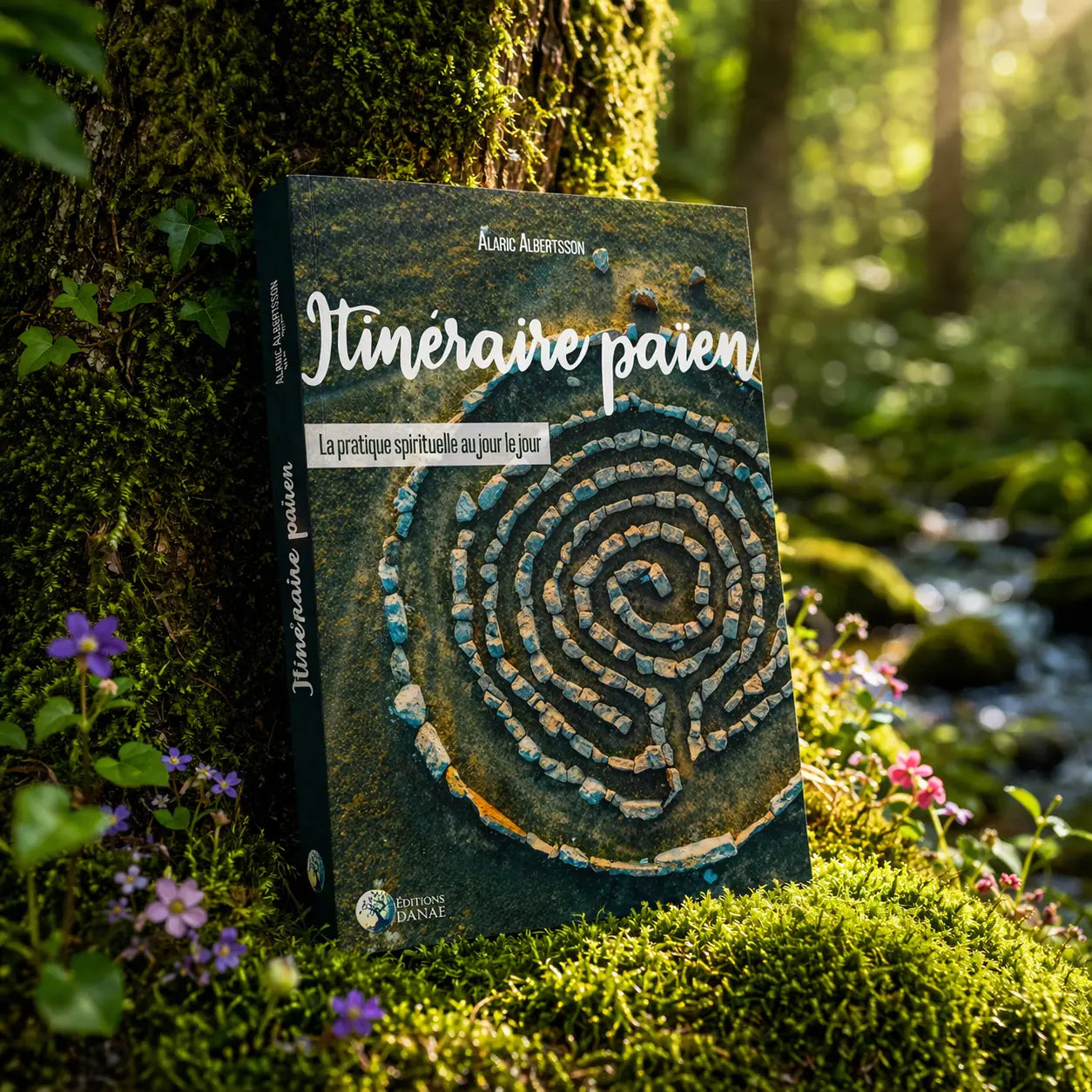 Livre Itinéraire Païen posé dans la mousse près d'un ruisseau, illustré d'une vue aérienne d'un labyrinthe de pierres sacrées au cœur de la nature.