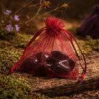 Pochette en organza bordeaux contenant plusieurs larmes d'apache polies d'un noir vitreux et profond, posée sur une branche d'arbre moussue en pleine nature.