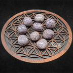 Neuf pierres de lépidolite roulées aux teintes mauves mouchetées et aux surfaces scintillantes, disposées sur un plateau en bois sculpté du motif de la "Fleur de Vie".