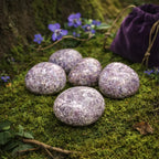 Cinq pierres de lépidolite polies aux nuances violettes et mauves irisées, disposées sur un tapis de mousse verte avec un pochon de velours violet en arrière-plan.
