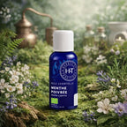 Flacon bleu d'huile essentielle de Menthe Poivrée Herbes et Traditions posé sur un tapis de mousse verte avec des fleurs blanches et un alambic en arrière-plan.