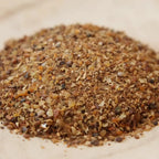 Gros plan sur un monticule de Myrrhe concassée, montrant un mélange de petits grains irréguliers et de poudre, de couleur brun-rouille et ambrée, sur une surface en bois clair.