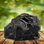 Bloc d'obsidienne noire brute à la cassure conchoïdale brillante, posé sur un plateau en bois devant un arrière-plan de feuillage vert flou.