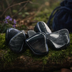 Plusieurs pierres d'obsidienne argentée polies, noires avec des reflets gris pailletés, disposées sur un tronc moussue avec un pochon bleu en arrière-plan.