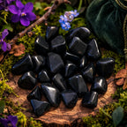 Amoncellement de pierres d'onyx noir polies, d'un noir profond et opaque, disposées sur un socle en bois moussue entouré de fleurs violettes et d'un pochon vert.