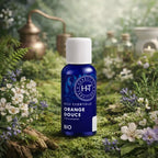 Flacon en verre bleu Herbes et Traditions - Huile Essentielle - Orange Douce Bio entouré de petites fleurs blanches sur un lit de mousse avec un alambic en arrière-plan.