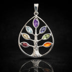 Un pendentif arbre de vie en argent en forme de goutte, dont les feuilles sont serties de sept pierres naturelles aux couleurs des chakras, présenté avec son reflet sur un fond noir.