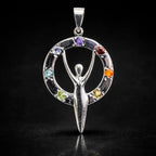 Un pendentif déesse en argent entourée d'un cercle serti de sept pierres naturelles aux couleurs des chakras, surmonté d'une bélière et présenté avec son reflet sur un fond noir.